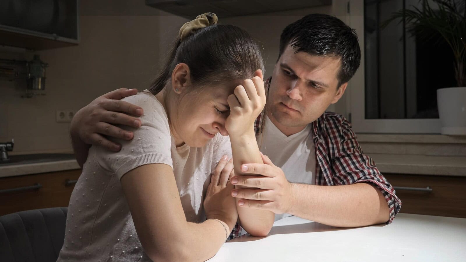 Young man consoling and hugging his crying wife on kitchen at night. Concept of despair, stress