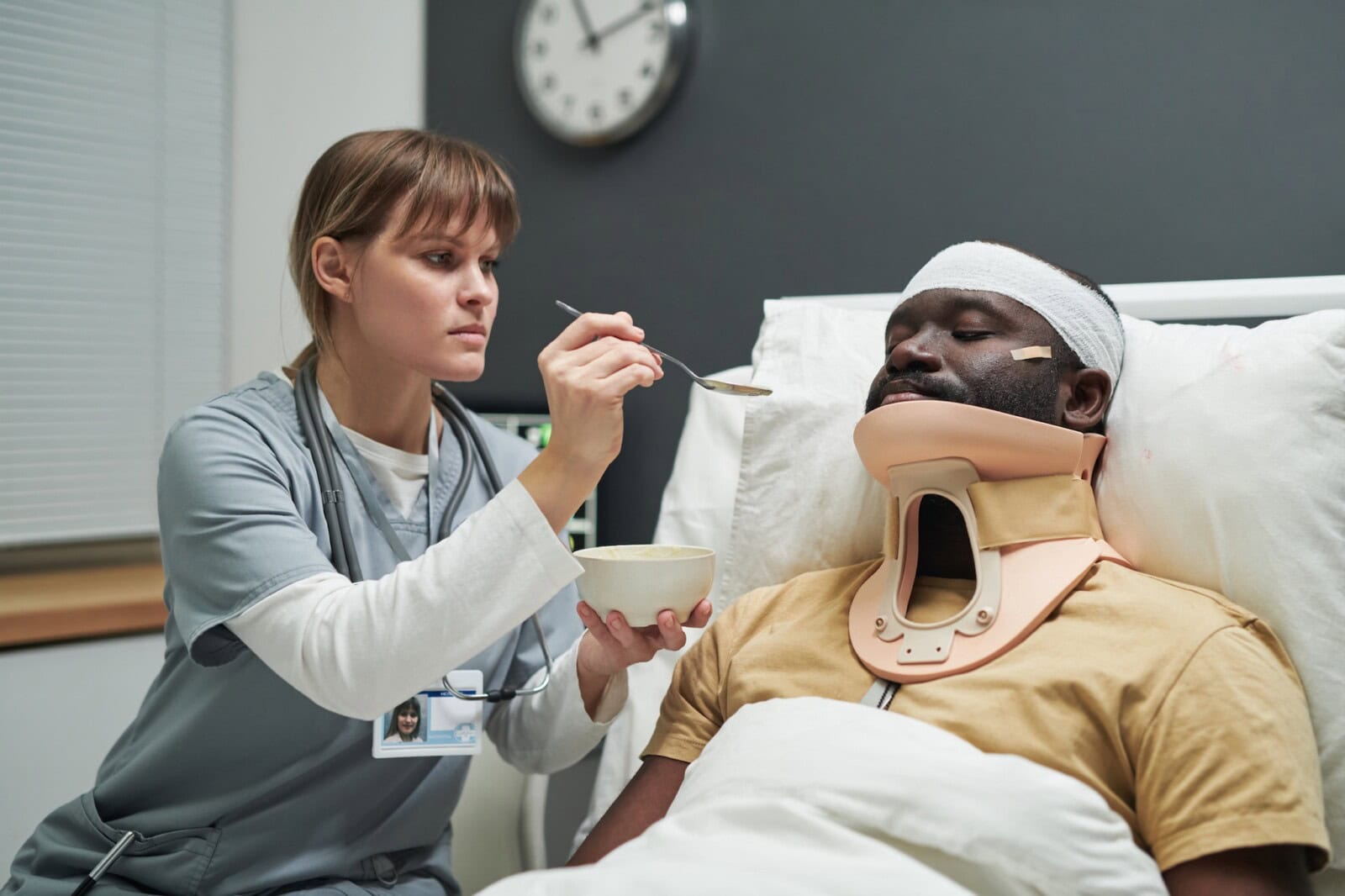 Nurse in uniform holding bowl with porridge while feeding injured patient