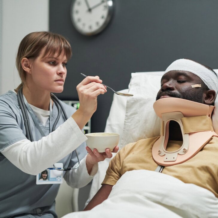 Nurse in uniform holding bowl with porridge while feeding injured patient